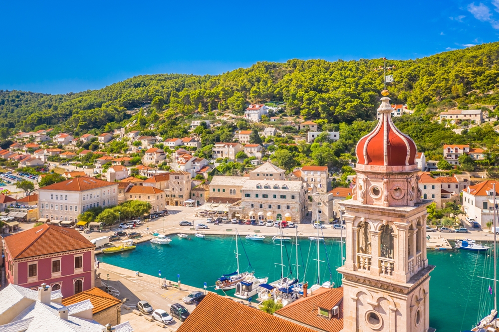 A shot of Croatia shows its natural greenery and tall buildings surrounding marinas, the peaked churches and sailboats contrasting each other and drawing attention. 