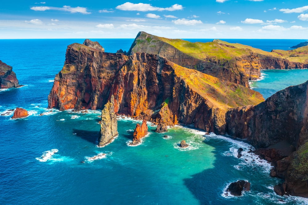 An arial shot of Madeira shows mountains, cliffs, and white capped waves as they crash against rocks in this stunning island. 