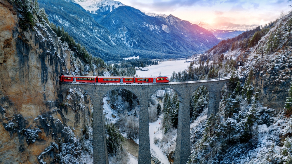 When considering which of the most iconic Europe Trips to Take in 2026, look into train travel through the Alps: here a red train travels from peak to peak of snowy mountains as the sun dips beyond them. 
