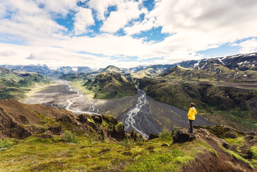 A woman in a yellow coat stands on the hugged landscape of the highlands in Iceland, looking over the mountains and rivers below. 