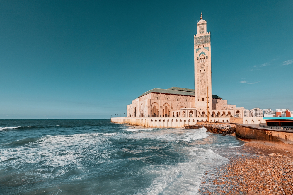 A coastal photo of Morocco shows why this is a iconic World Trips to Take in 2026: the blue water bounces in white capped waves on the sore and beyond a temple that sits in the middle of the water on a pier. 