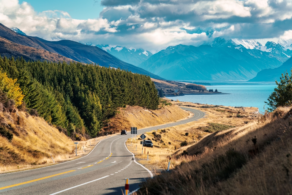 A winding road on the coast of New Zealand shows rugged landscapes, trees, mountains, rocky sides, and water. 